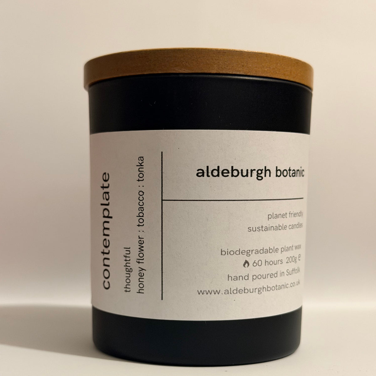 A matte black candle jar with a light brown wood-look lid and a white paper label. The label reads 'aldeburgh botanic' and 'contemplate' with scent text 'honey flower : tobacco : tonka'.