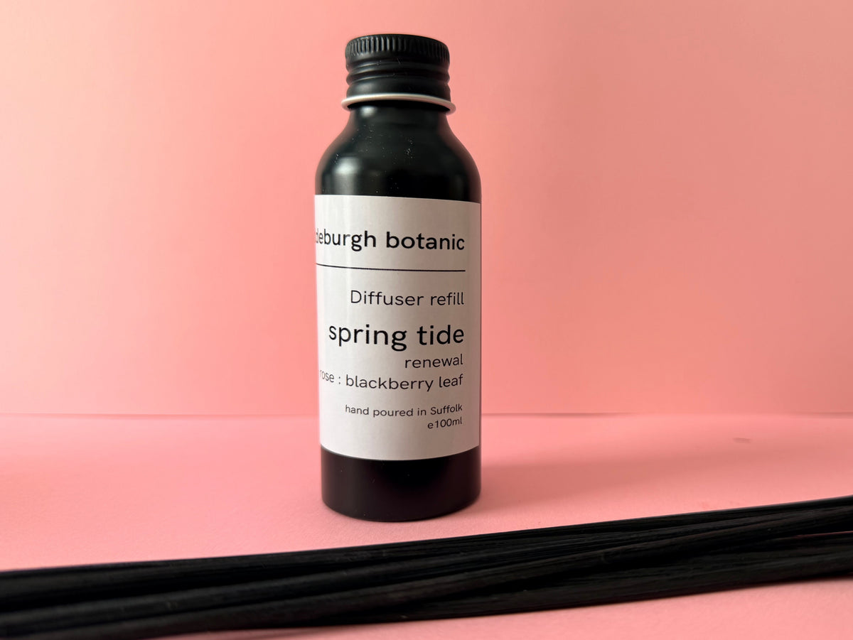 A black screw top aluminium bottle with white label of Aldeburgh diffuser oil refill with black diffuser reeds laying in front, shown against a pink background.
