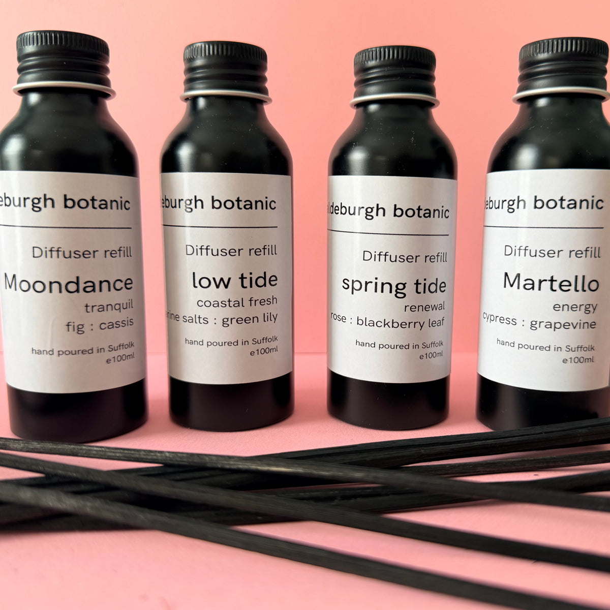 Four black screw top aluminium diffuser refill bottles with white labels from Aldeburgh Botanic, arranged on a pink background with several black reeds in the foreground. The labels read: Moondance, low tide, spring tide, and Martello.